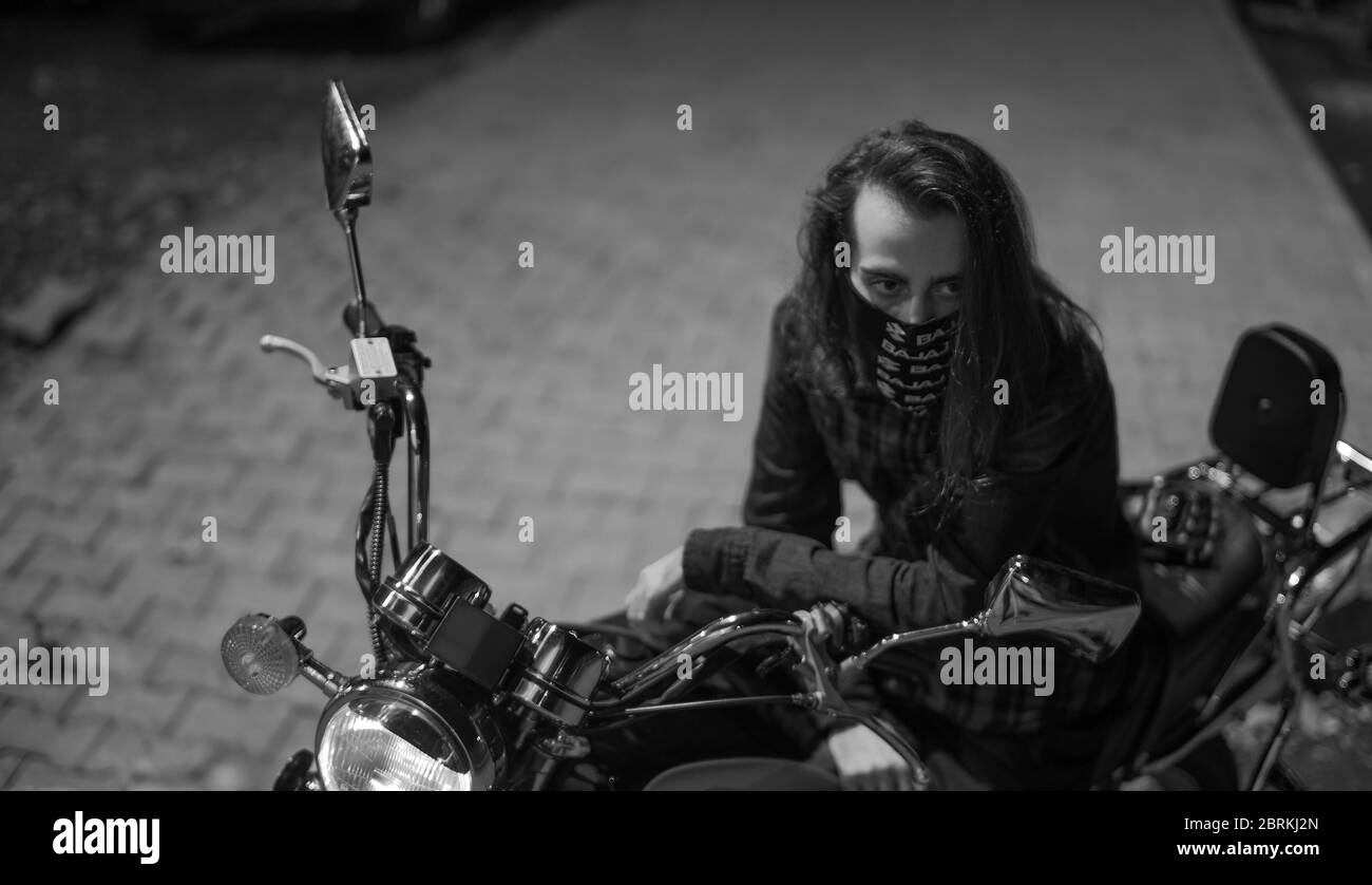 Young man with long hair mask on motorcycle on night street. Black and white color Stock Photo