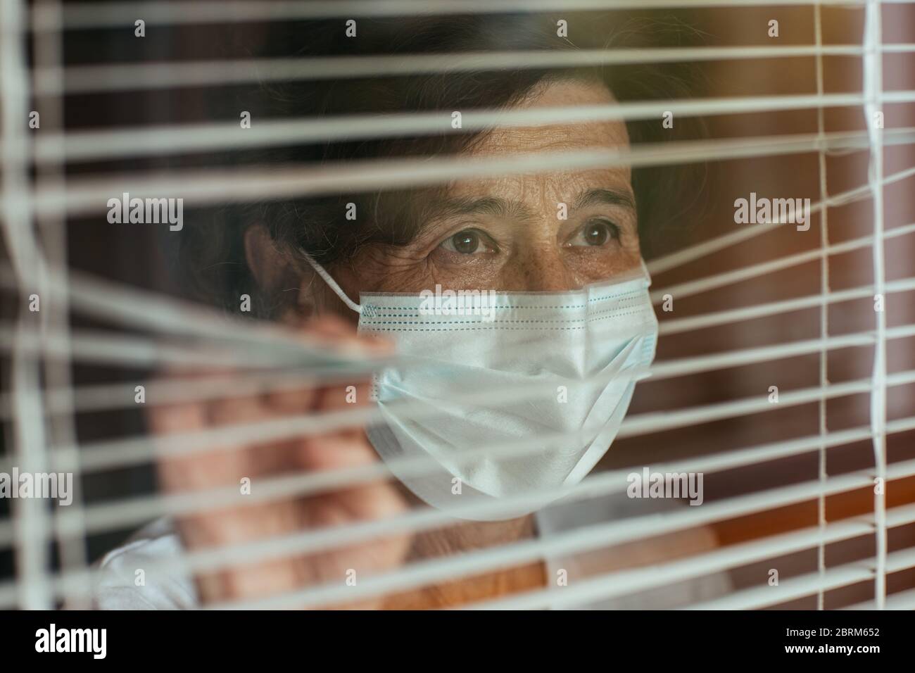 Close up of anxious old woman surgical mask looking through window blinds during self-quarantine. Senior female wearing face mask. Stock Photo