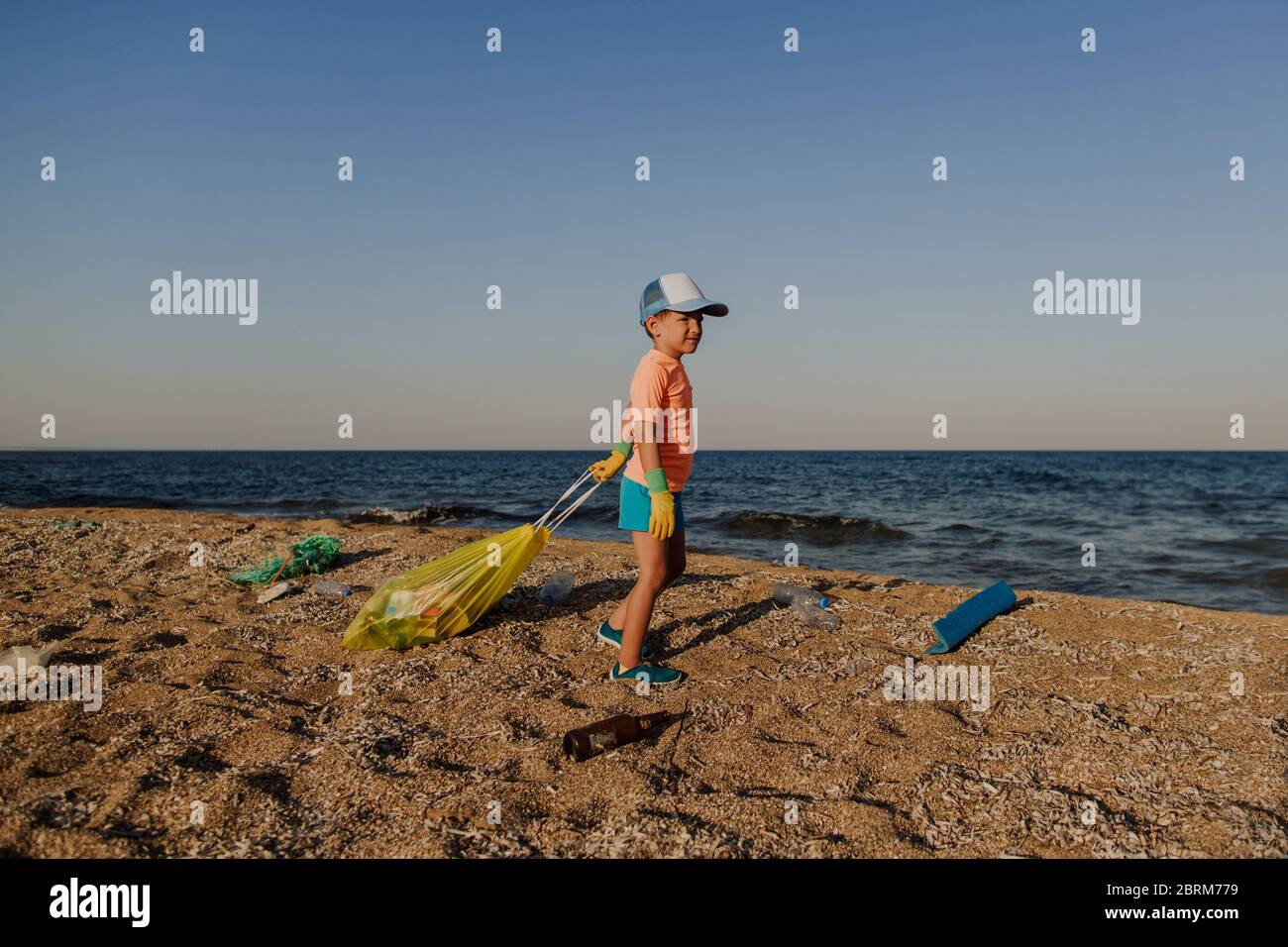 Boy cleaning beach with bag full of rubbish. 5 year old child wearing rubber gloves taking responsibility for the environment and collecting waste. Stock Photo