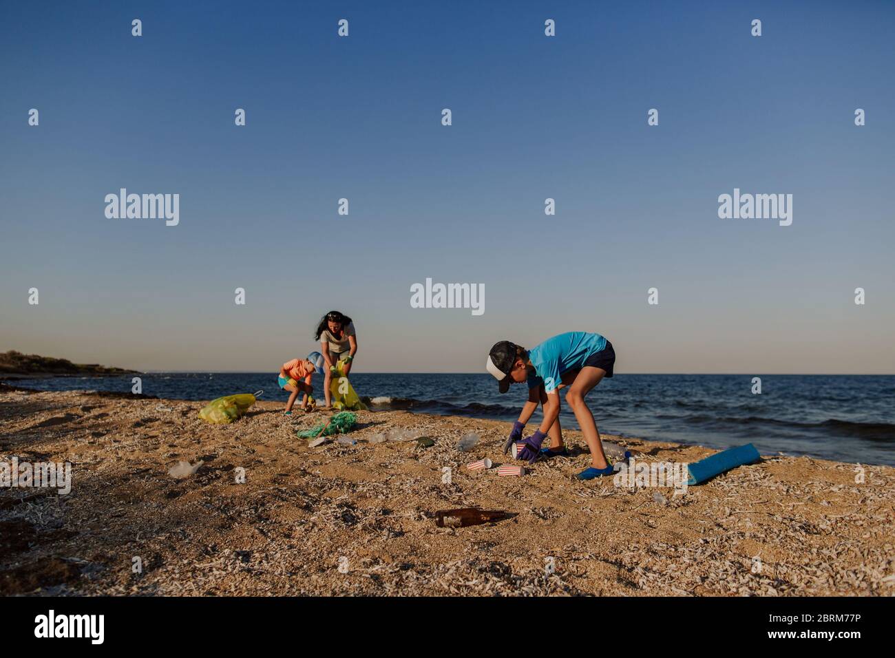 Family picking up rubbish on polluted beach. Mother and two sons cleaning beach and putting garbage into plastic bin bags. Stock Photo