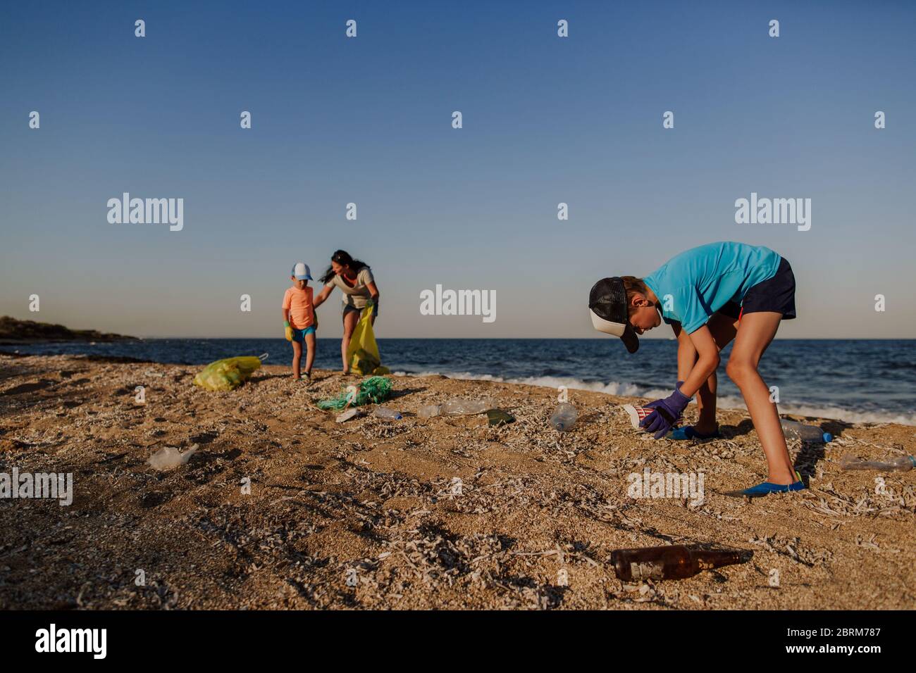 Boy collecting litter on beach with his mother and brother. Two boys and woman picking up garbage found on the beach and putting it into plastic bin. Stock Photo