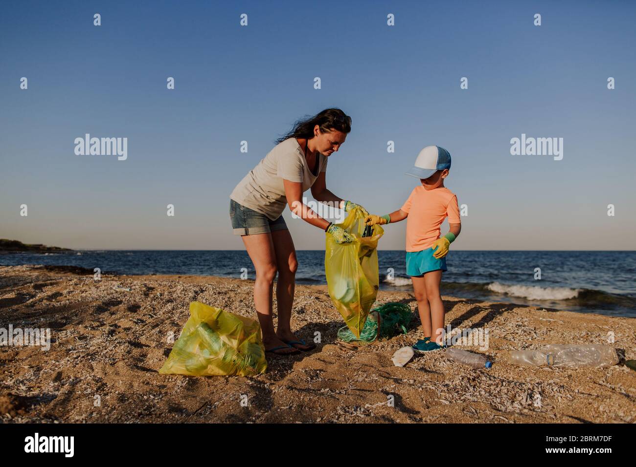 Mother collecting rubbish with son on beach. Woman and boy wearing rubber gloves filling plastic bag with rubbish left on the beach. Stock Photo