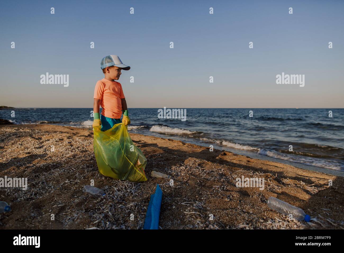 5 year old boy standing on beach holding bin bag collecting litter. Child wearing rubber gloves
