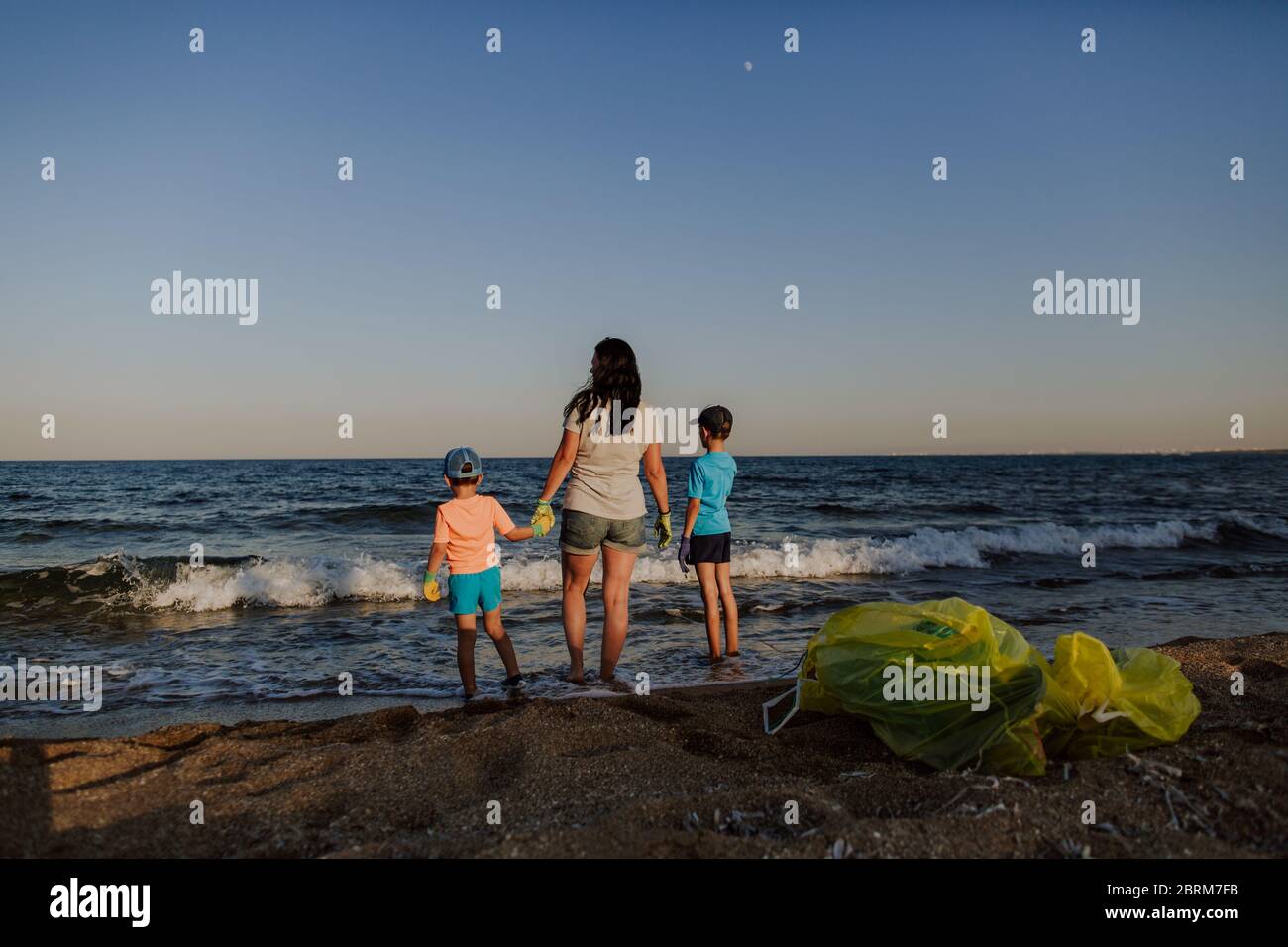 Mother and two sons standing on beach with rubbish bags on shore. Woman and two boys wearing rubber gloves looking out to sea with breaking waves. Stock Photo