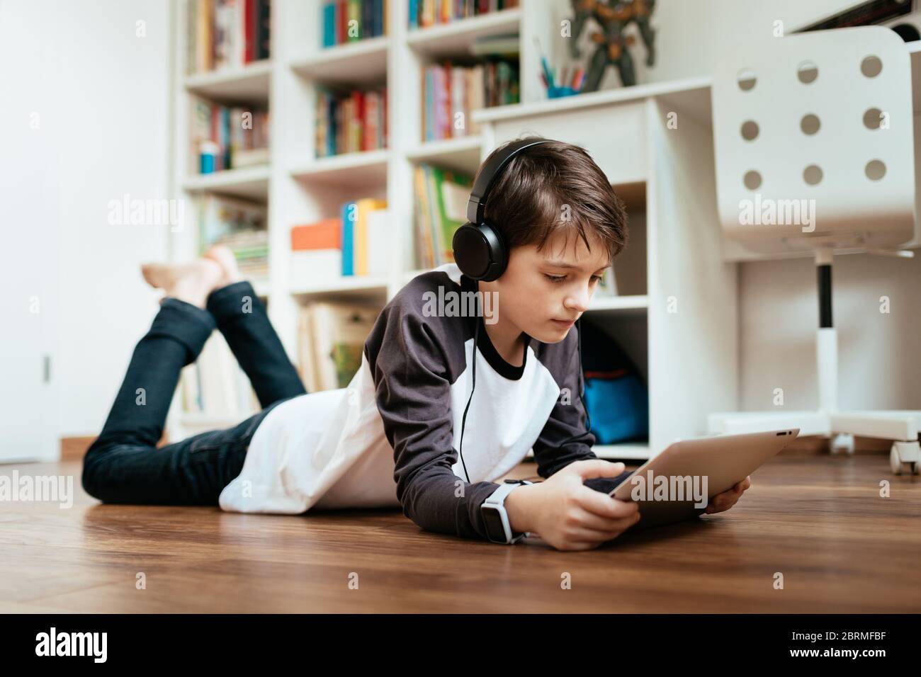 Young student wearing headphones lying on the floor studying at home working with digital tablet. Boy relaxing and online learning on digital tablet. Stock Photo