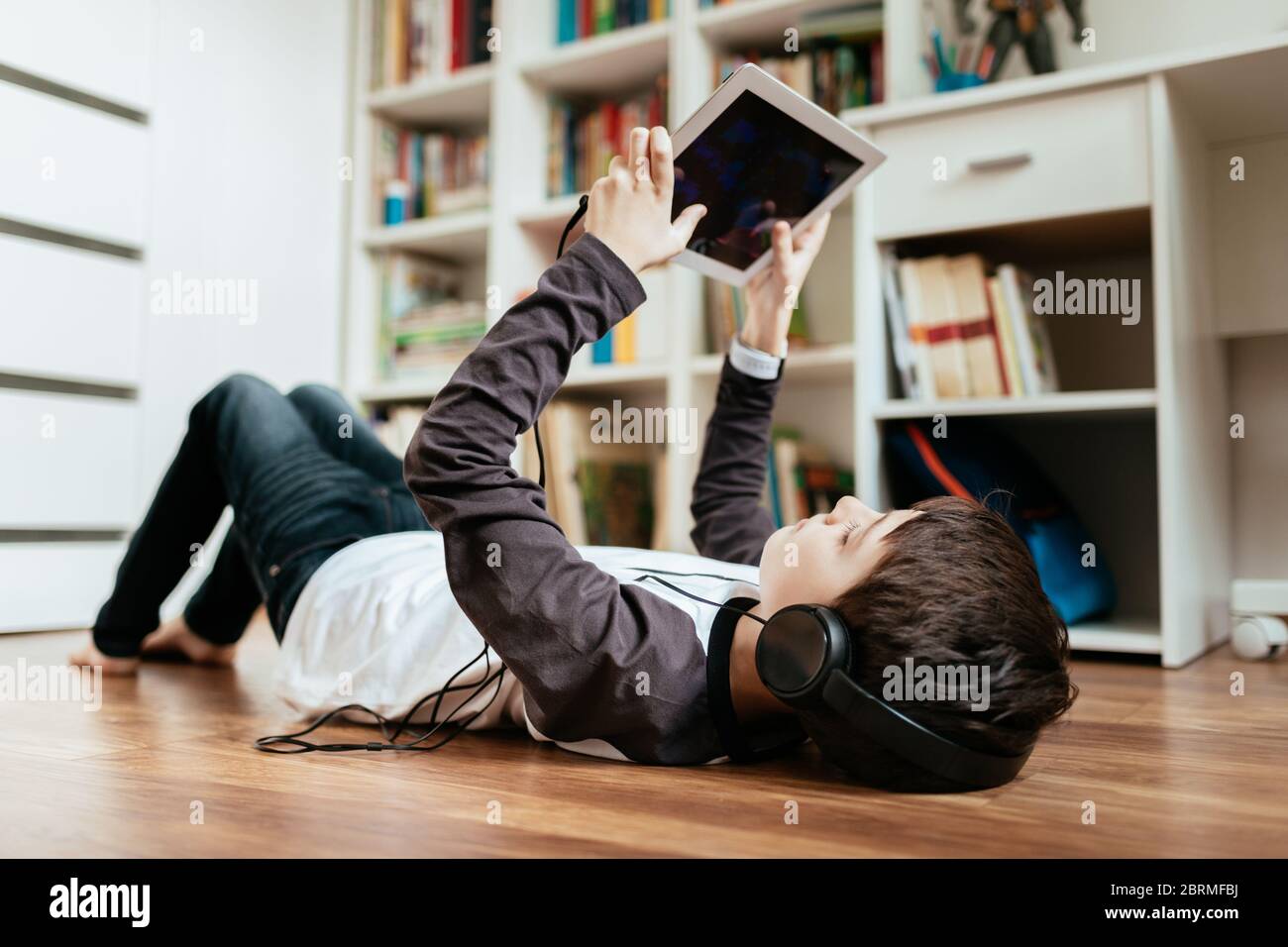 Teenage boy wearing headphones lying on the floor playing game on tablet at home. Boy relaxing by playing online game on digital tablet. Stock Photo