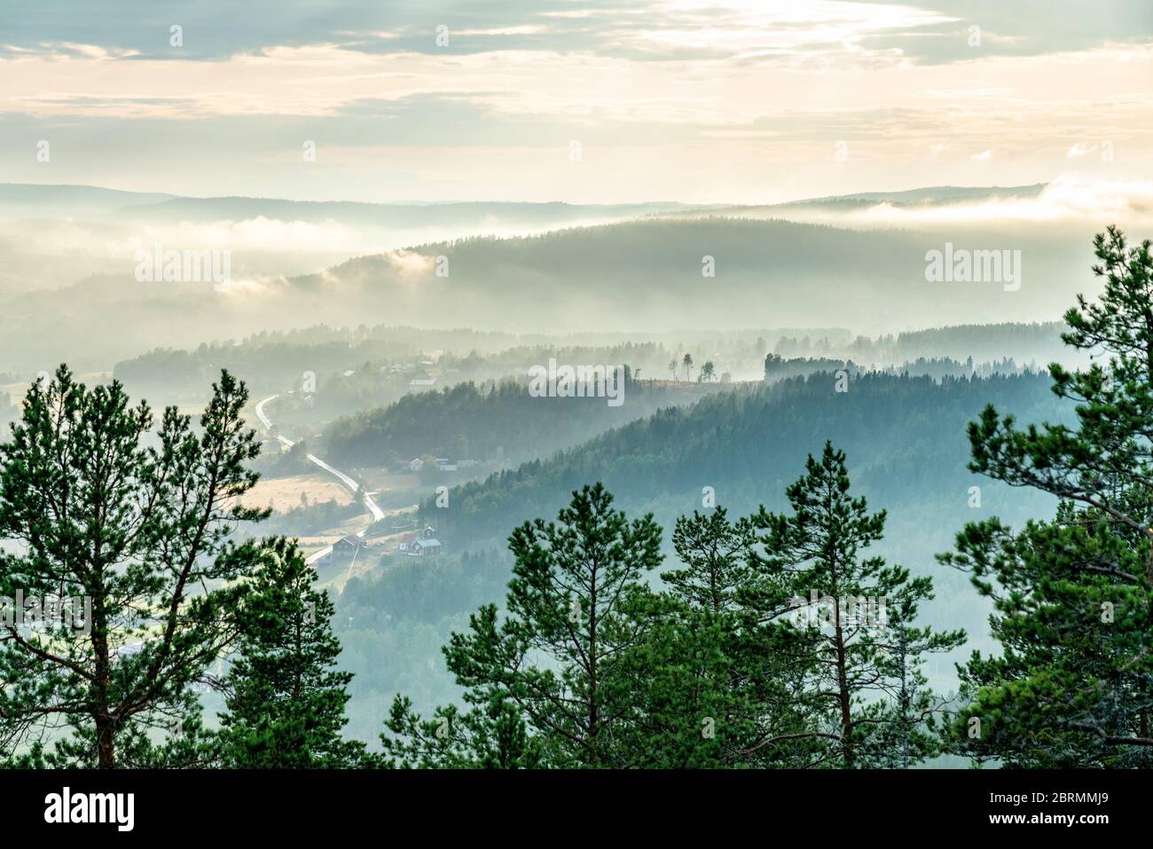 View from the mountain top over foggy Scandinavian mountains with wild pine tree forest, the village, mountain ridges, summer day with heavy dramatic Stock Photo