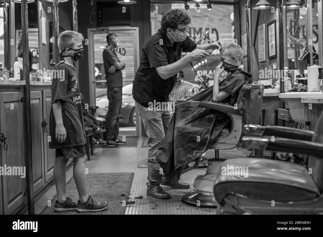 May 20, 2020, Manhattan, Kansas, USA: DYLAN ROBERTS, center, cuts CLARK WEDDLE'S, 10, hair on Wednesday as LANCE MULLET, left, watches. MULLET, has been staying with the WEDDLE'S as his mother works long hours as a custodian at a local Church. Campus Barbershop in Aggieville, Kansas's oldest shopping district, has been open for two days after being closed since March, 17. May, 18, was the first day of Phase 1.5 of the plan to reopen Kansas's economy. ROBERTS, restricting the number of patrons inside the barbershop did allow the family to come in together. (Credit Image: © Luke Townsend/ZUMA Wi Stock Photo