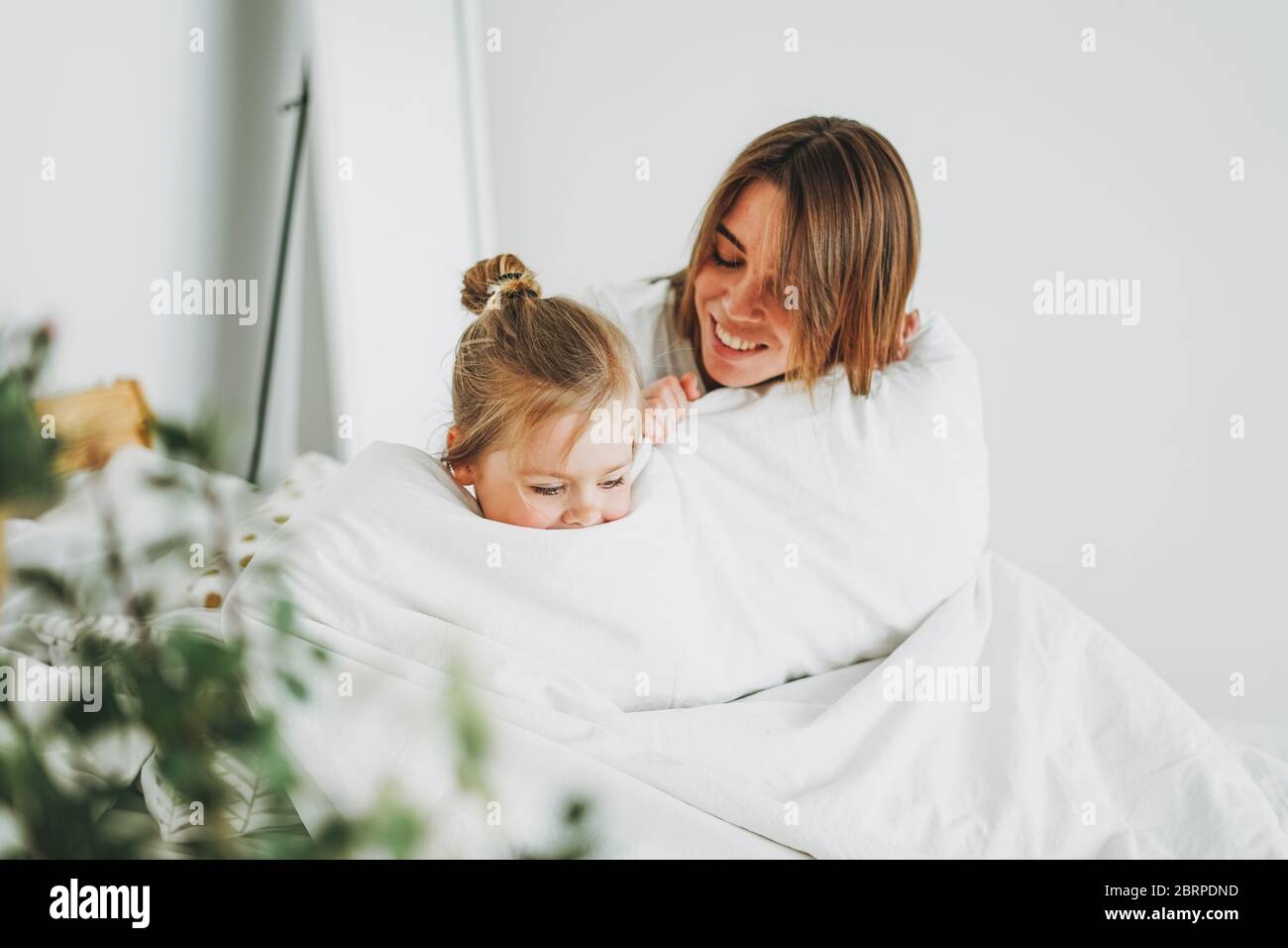 Cute toddler girl long fair hair big grey eyes looking at camera having fun with mother on bed stay at home Stock Photo