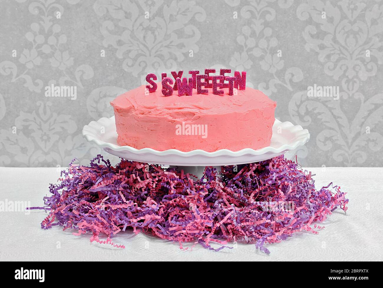 A pretty pink birthday layer cake with Sweet Sixteen spelled out on top in glitter candles.  Cake is on pedestal and surrounded by pink and purple cri Stock Photo
