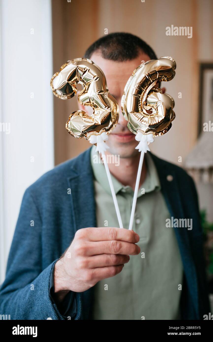 man in a blue jacket holds in his hands little foiled gold balloons of the number 36 on sticks and covers his face with them. Thirty-sixth birthday. T Stock Photo