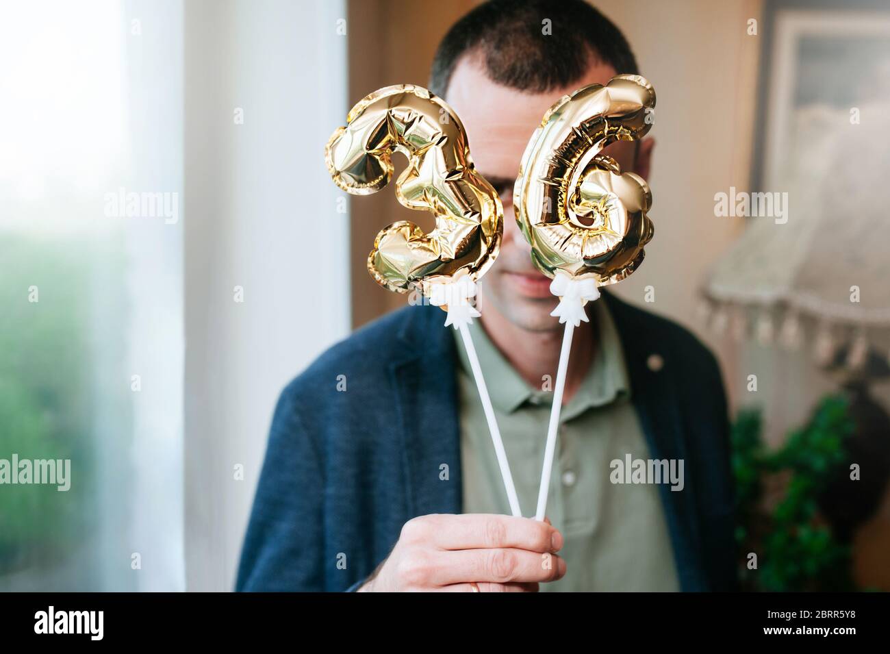 man in a blue jacket holds in his hands little foiled gold balloons of the number 36 on sticks and covers his face with them. Thirty-sixth birthday. T Stock Photo