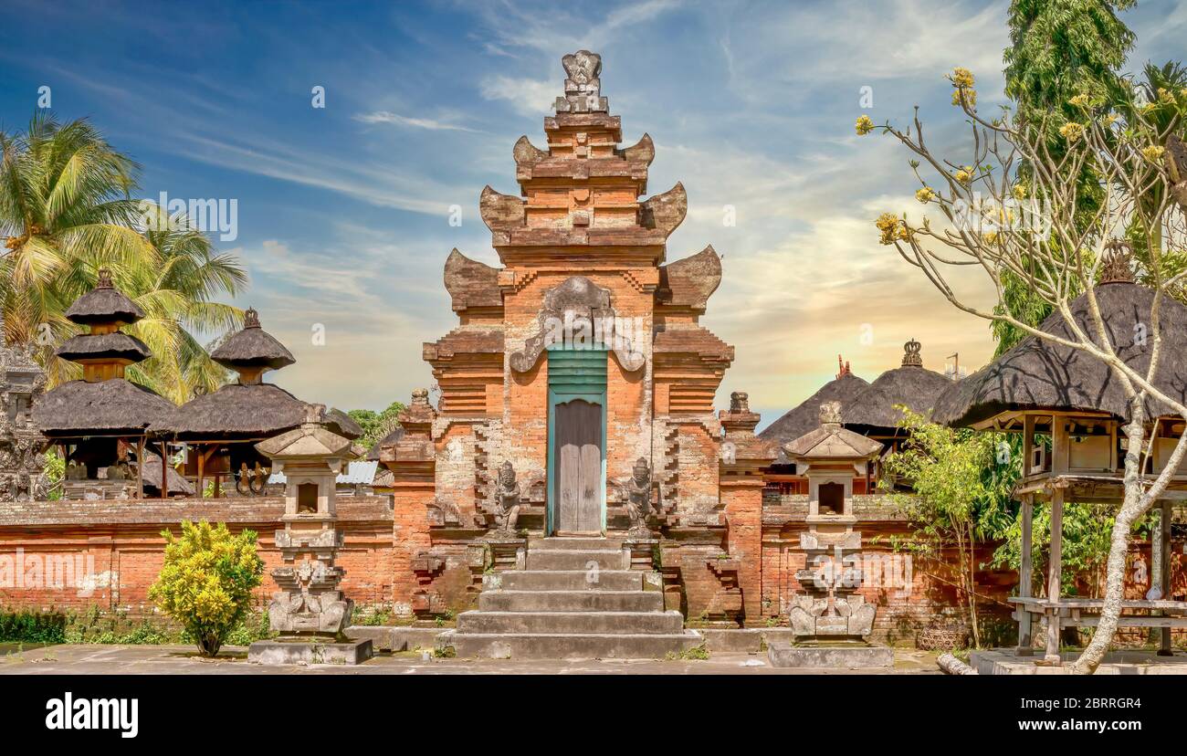 Street view of a Balinese Hindu temple complex near Ubud, with a red brick tower gate and exterior wall, and thatched roofs of interior pavilions. Stock Photo