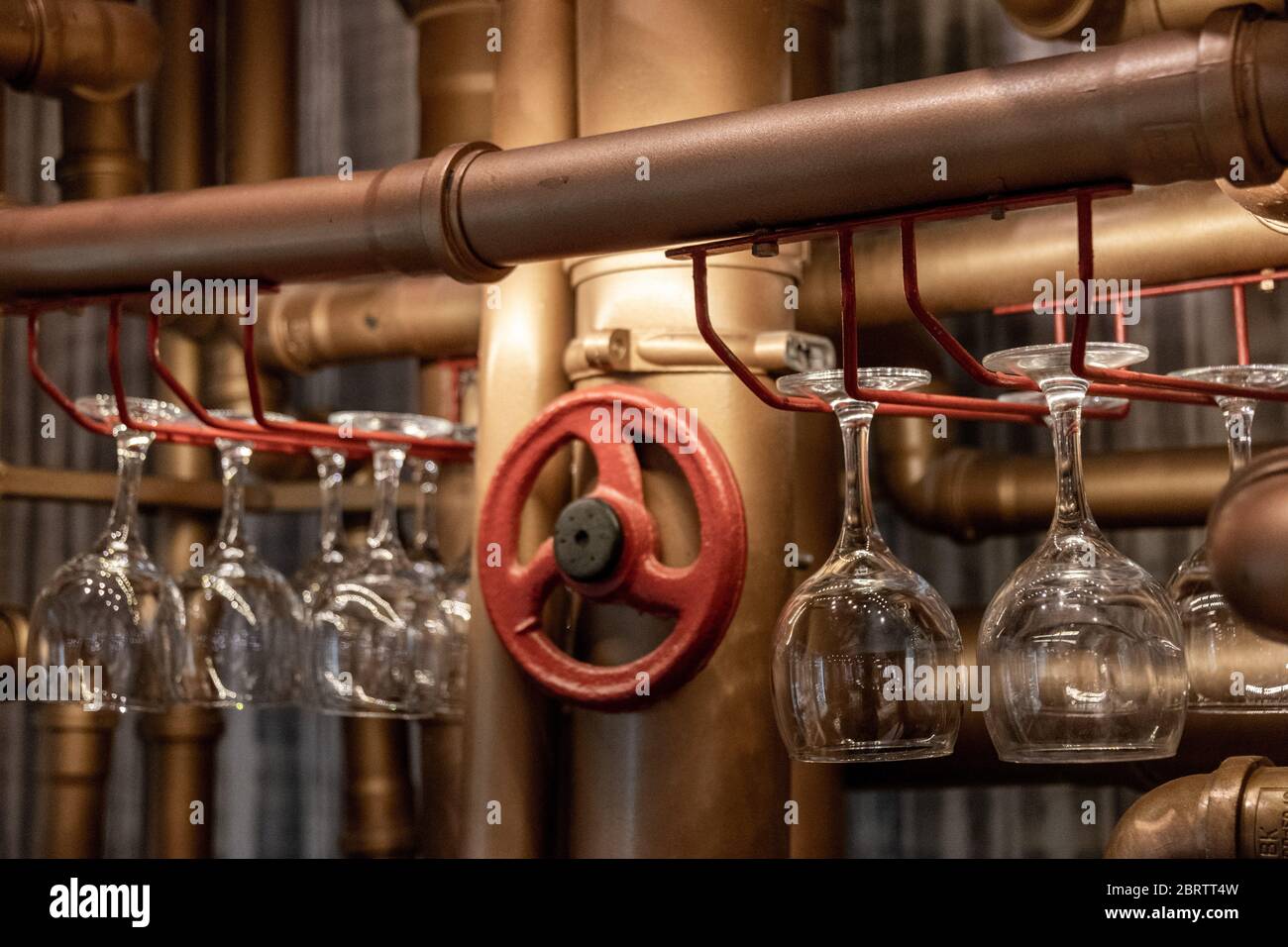 Shiny transparent empty wineglasses are hanging upside down on bar shelves made from water pipes painted in bronze and red colors. Drinking glasses di Stock Photo