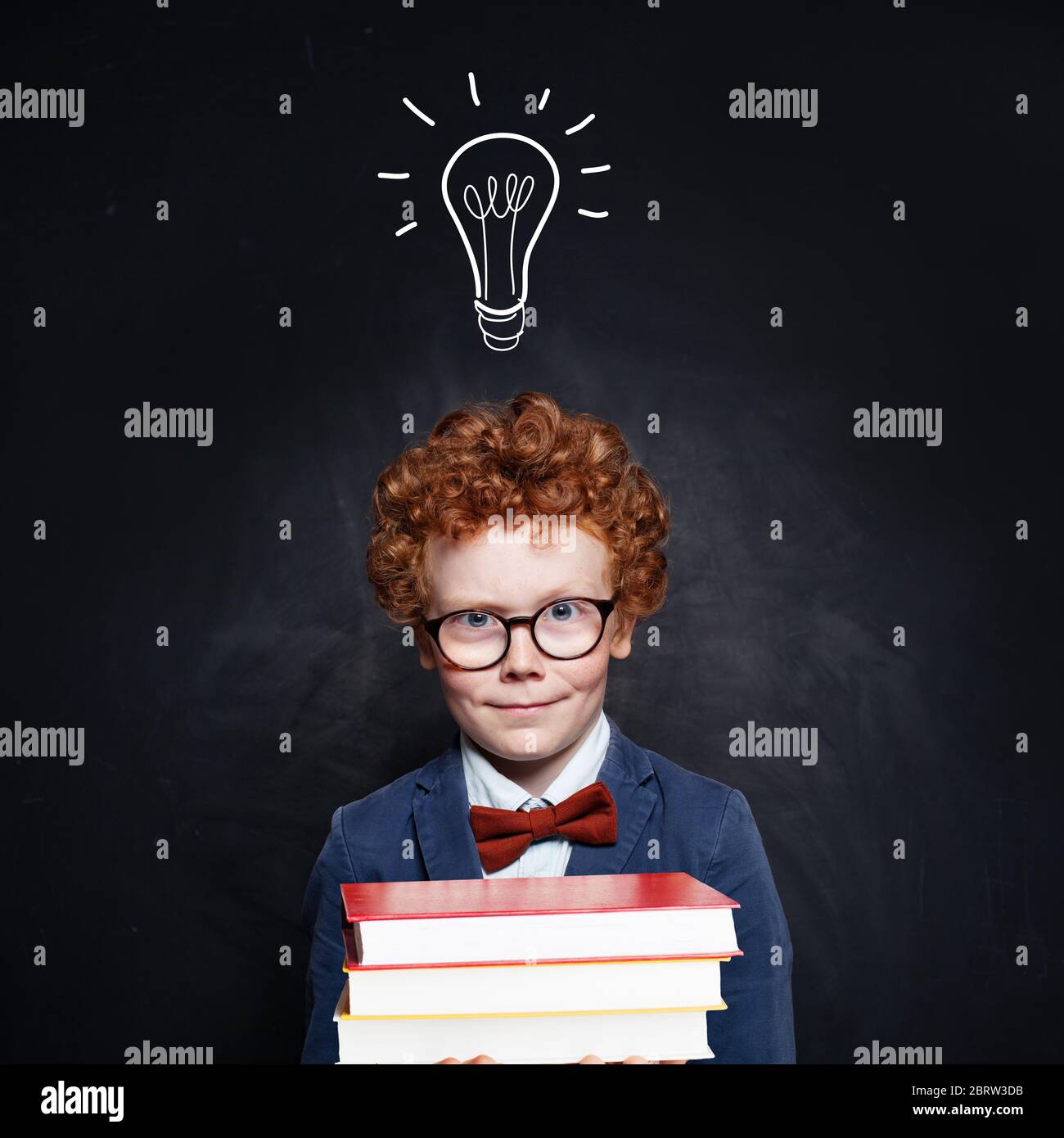 Happy kid with ginger hair holding stacks of book on school blackboard background with idea lightbulb Stock Photo