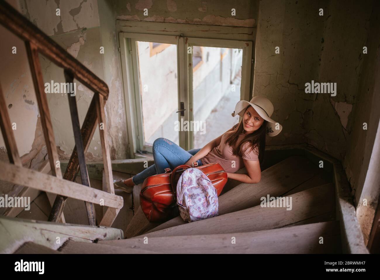 A beautiful young caucasian woman with a straw hat, backpack and a suitcase on the steps of an old abandoned building Stock Photo