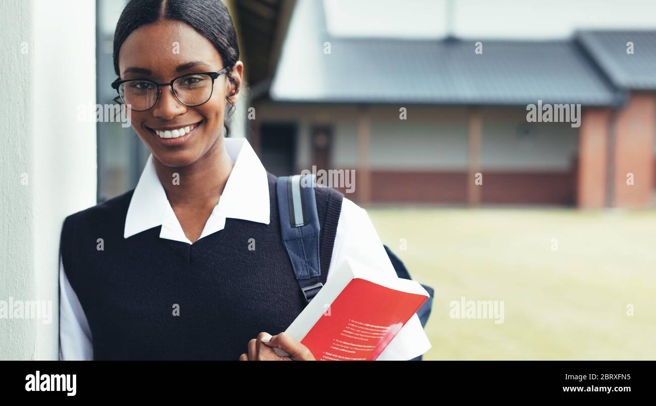 Teenage girl holding a textbook walking through the high school corridor. Confident high school student in uniform looking at camera and smiling, goin Stock Photo