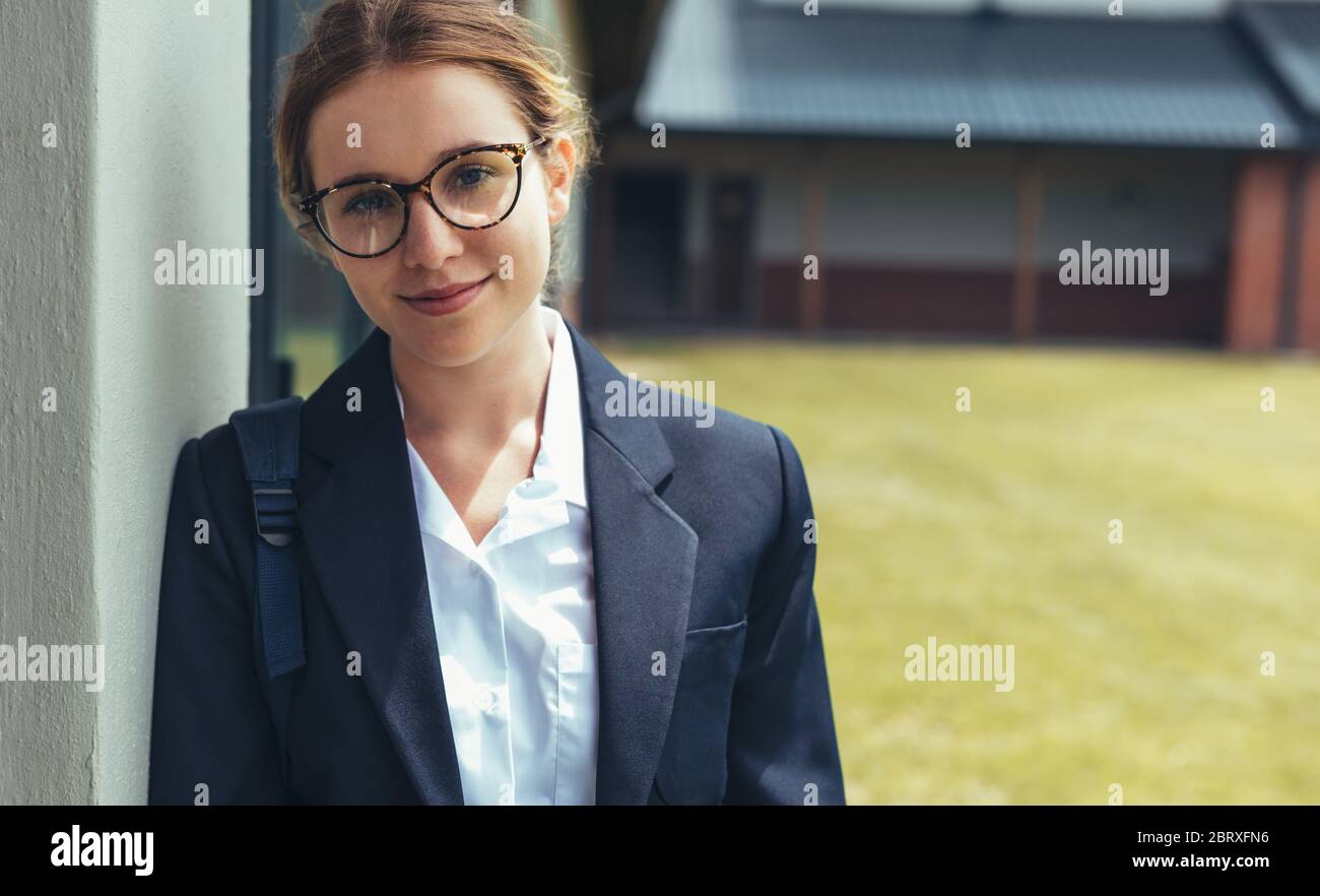 Teenage girl with schoolbag standing in high school campus. Female high school student in uniform looking at camera. Stock Photo