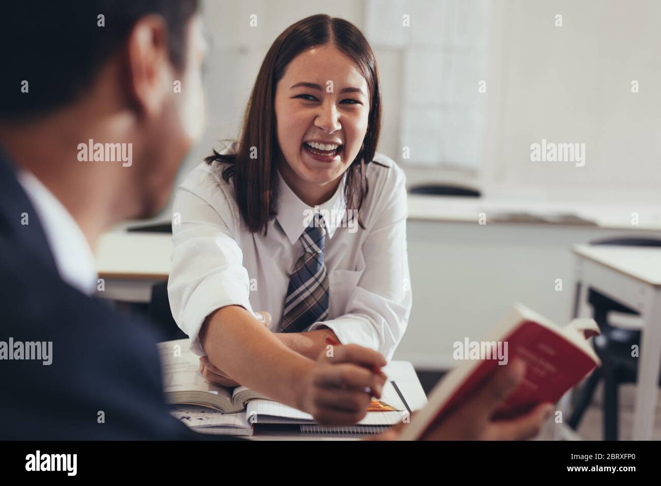 Two students talking and smiling in classroom. Young girl talking with her classmate in high school classroom. Stock Photo