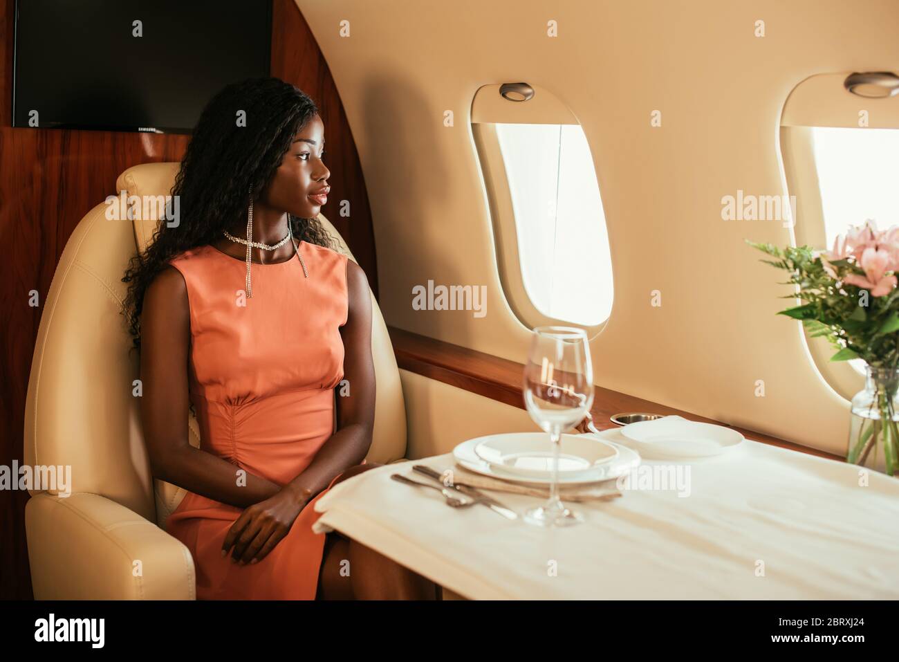 attractive, elegant african american woman looking in porthole of private plane while sitting at served table Stock Photo