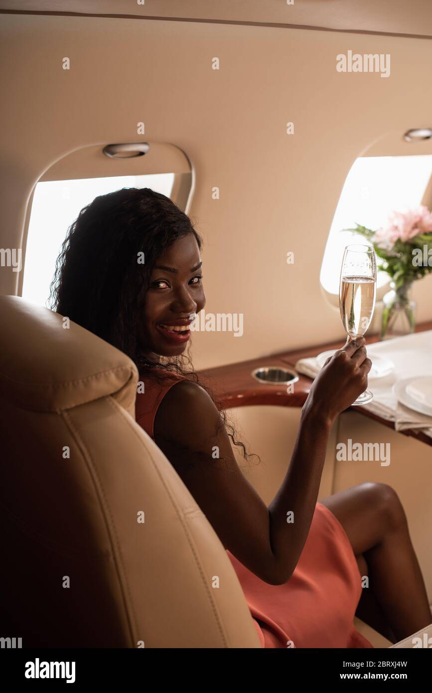 attractive african american woman travelling in private jet, holding glass of champagne and smiling at camera Stock Photo