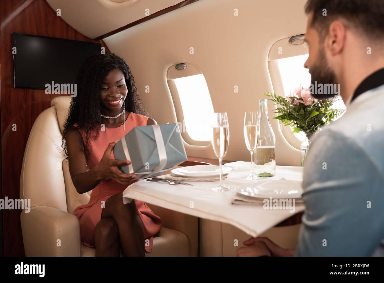 selective focus of happy african american girl holding gift box near man and served table in private plane Stock Photo