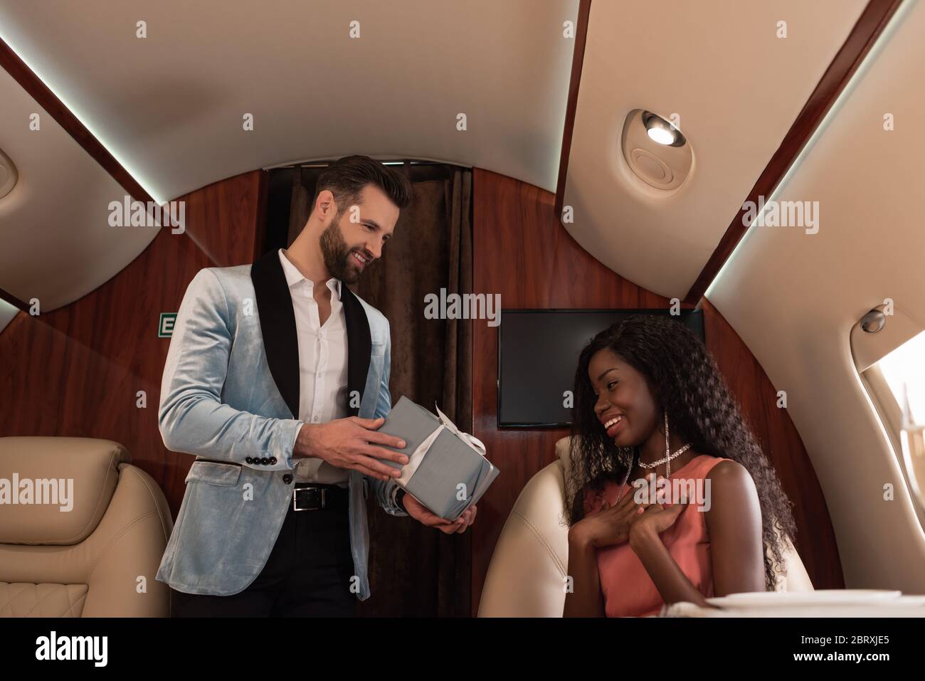 handsome, elegant man presenting gift box to suprised african american woman in private plane Stock Photo