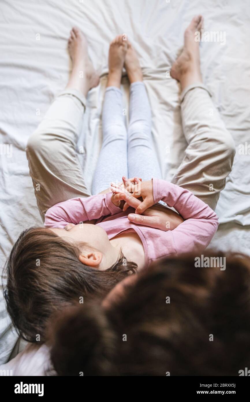 A little girl lays back on her mother's chest in the bed as they hold hands together. Mom wraps her in her arms as a sign of love Stock Photo