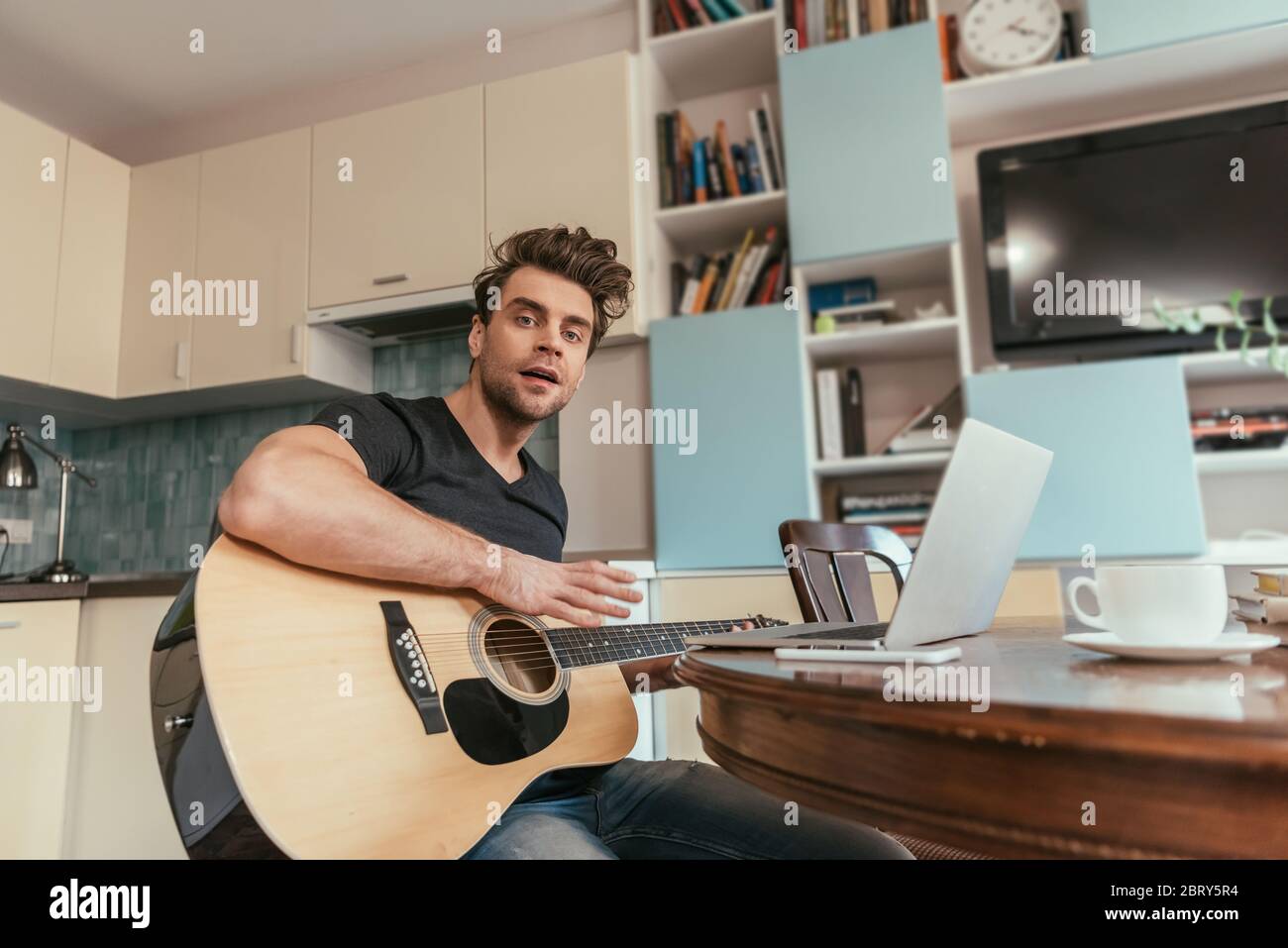 low angle view of handsome man holding guitar while sitting near laptop Stock Photo