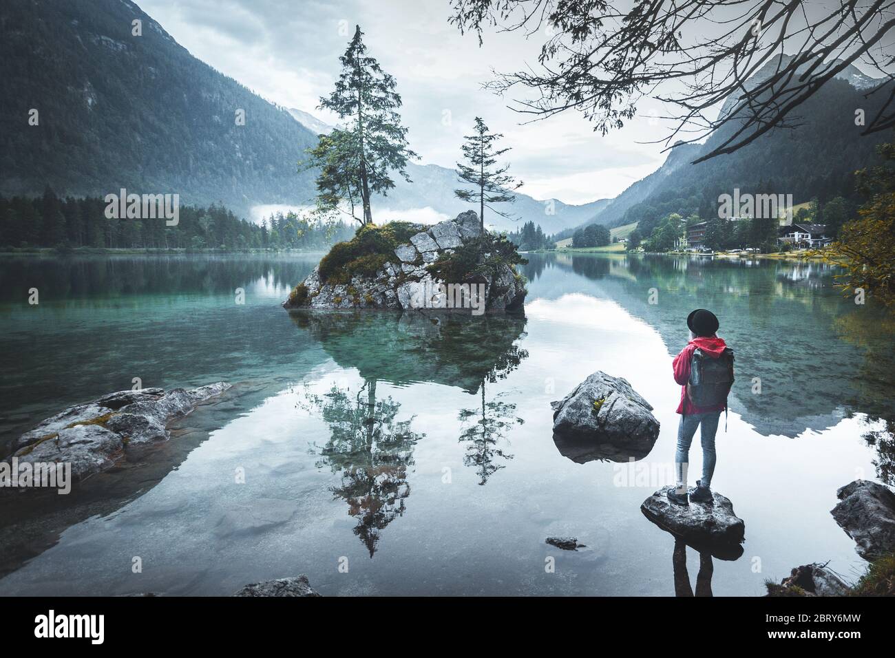 Scenic view of a beautiful woman standing on rock near lakem wearing a red jacket, a hat and a backpack looking at mountains, Lake Hintersee, Berchtes Stock Photo