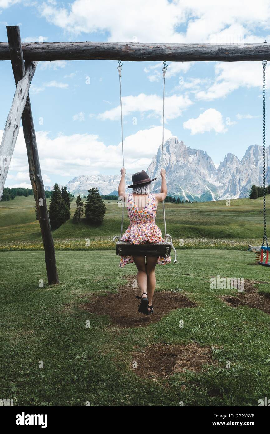 Back view of happy blonde girl on swing against blue sky in Seiser Alm, Dolomites, Italy. Carefree woman with floral dress and a hat enjoys the mounta Stock Photo