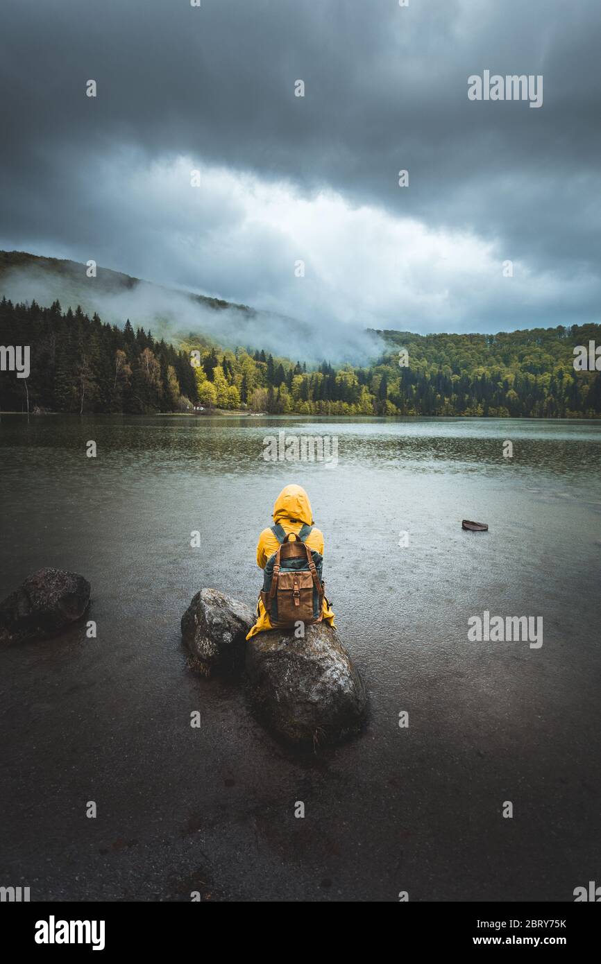 Amazing rear view reflection of a woman standing on rock in the middle of on a rainy summer day by the lake. Folk scenery concept with cloudy sky and Stock Photo