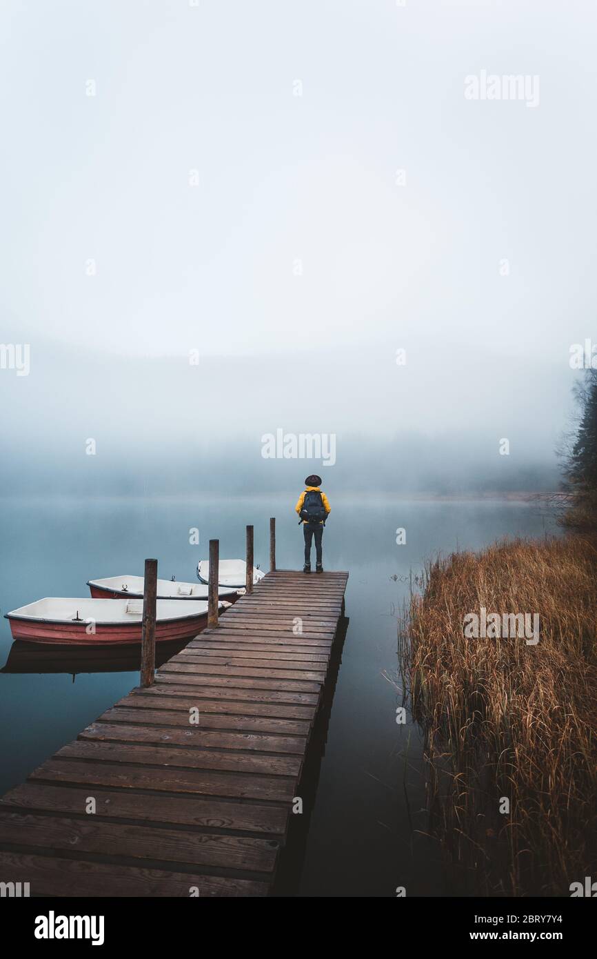 Backpacker standing on pier on a moody sunrise at Lake Sfanta Ana with anchored boats against foggy mountains Stock Photo