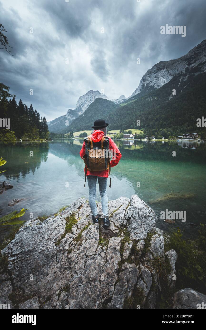 People and nature concept. Female tourist enjoys morning view from shore, looks at mountains and lake, being solo traveller, watching beauty, carries Stock Photo