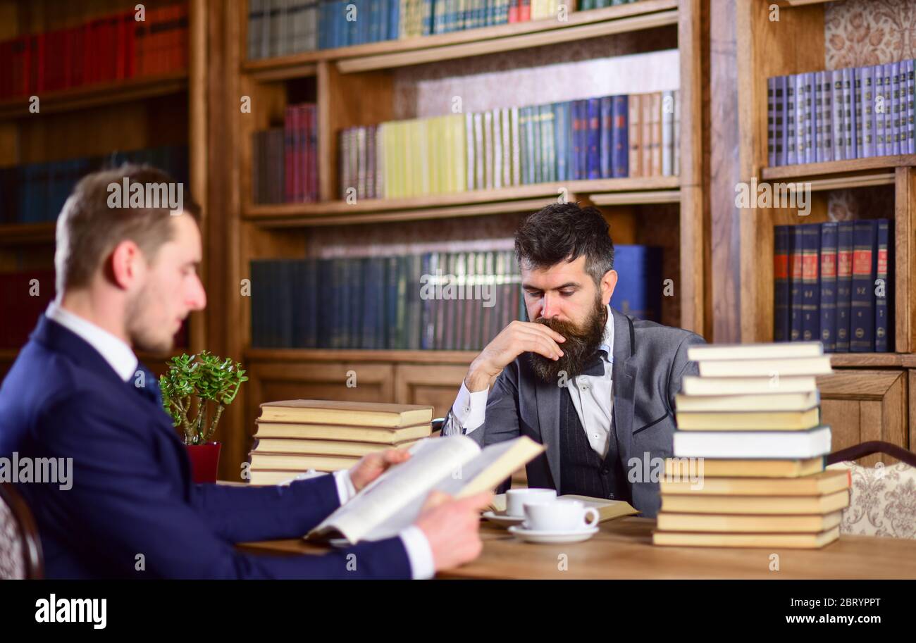 Intellectuals and aristocrats concept. Men in suits, aristocrats, professors in library or vintage interior with bookshelves on background. Men with busy faces reading books, studying and drink tea. Stock Photo