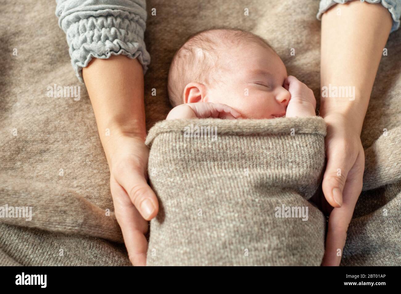 A four week old baby swaddled in a brown blanket, being held by her mother, asleep. Stock Photo