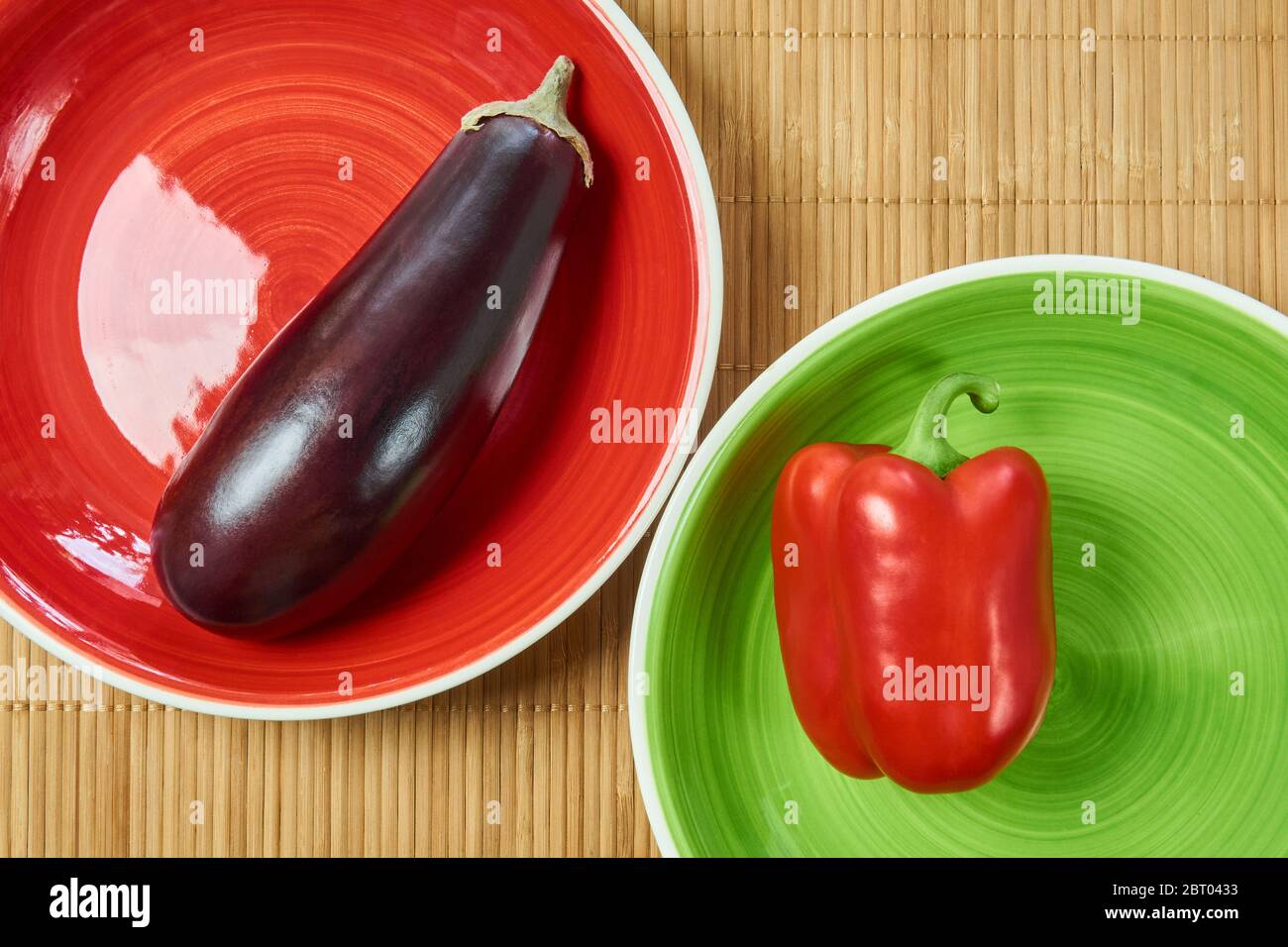 color contrasting still life - purple eggplant on a red plate and red bell pepper on a pale green plate next on a cane tablecloth Stock Photo