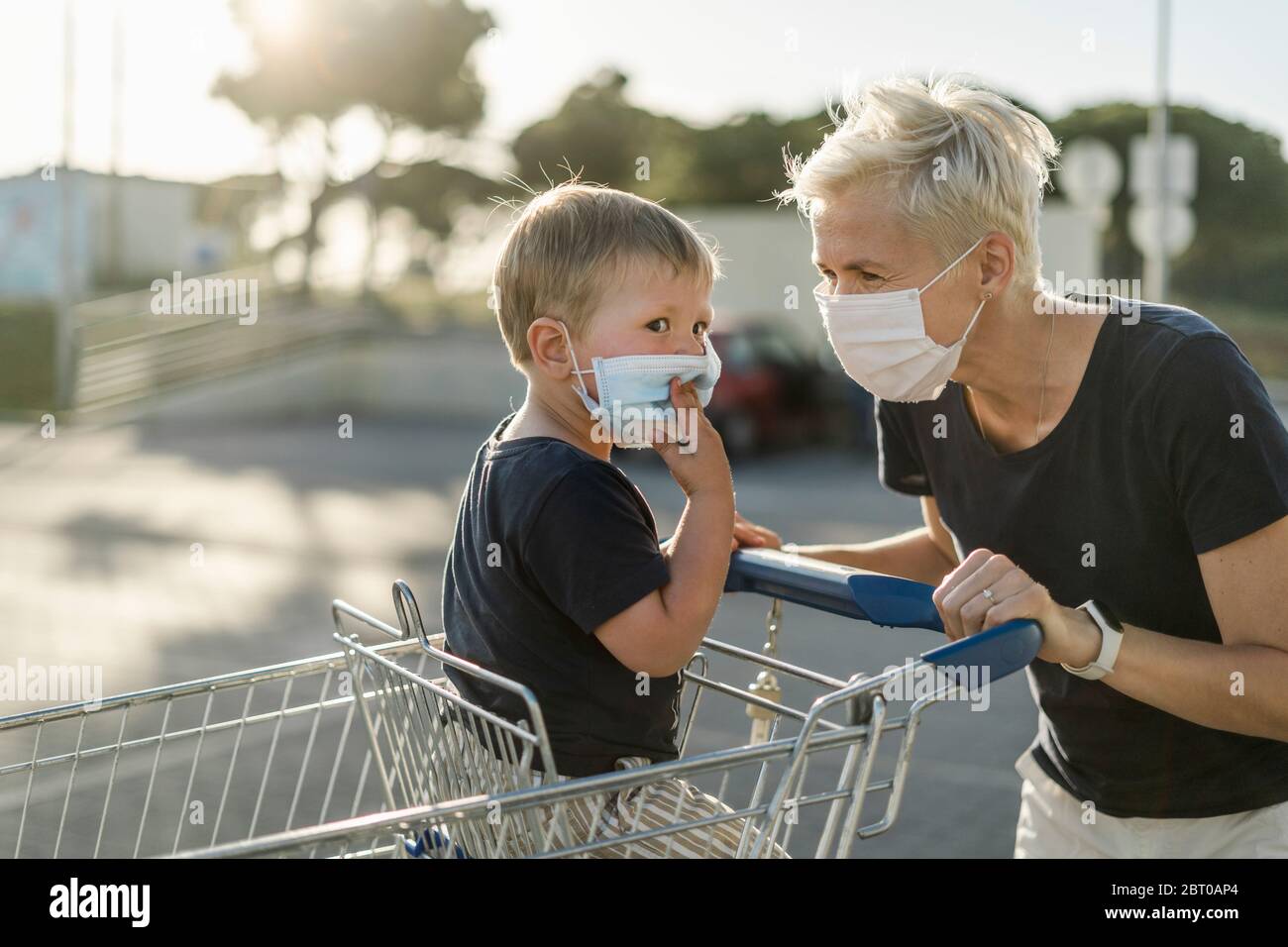 Mother joyfully playing with kid sitting in shopping cart. Both wearing protective face mask before entering a supermarket. Stock Photo