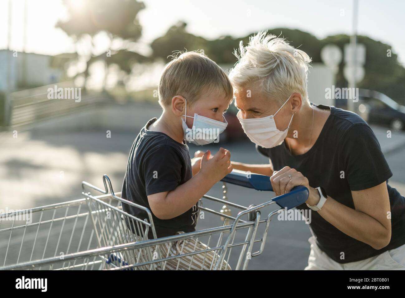 Mother joyfully playing with kid sitting in shopping cart. Both wearing protective face mask before entering a supermarket. Stock Photo