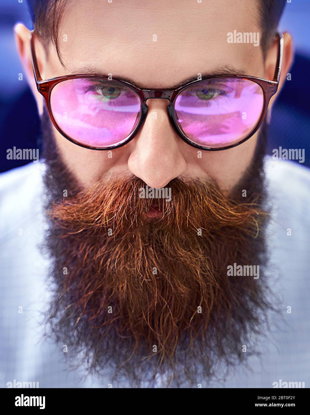 Bearded man in glasses with screen reflection working on computer in IT office, sitting at desk writing code, working on a project in software Stock Photo