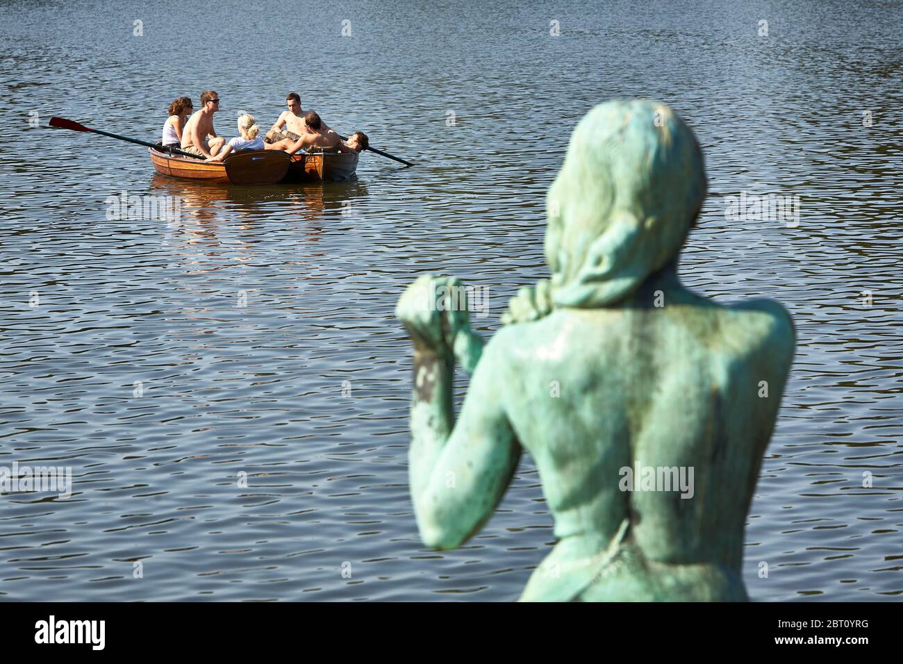 A coppery female statue arranges her hair while youngsters in rowing