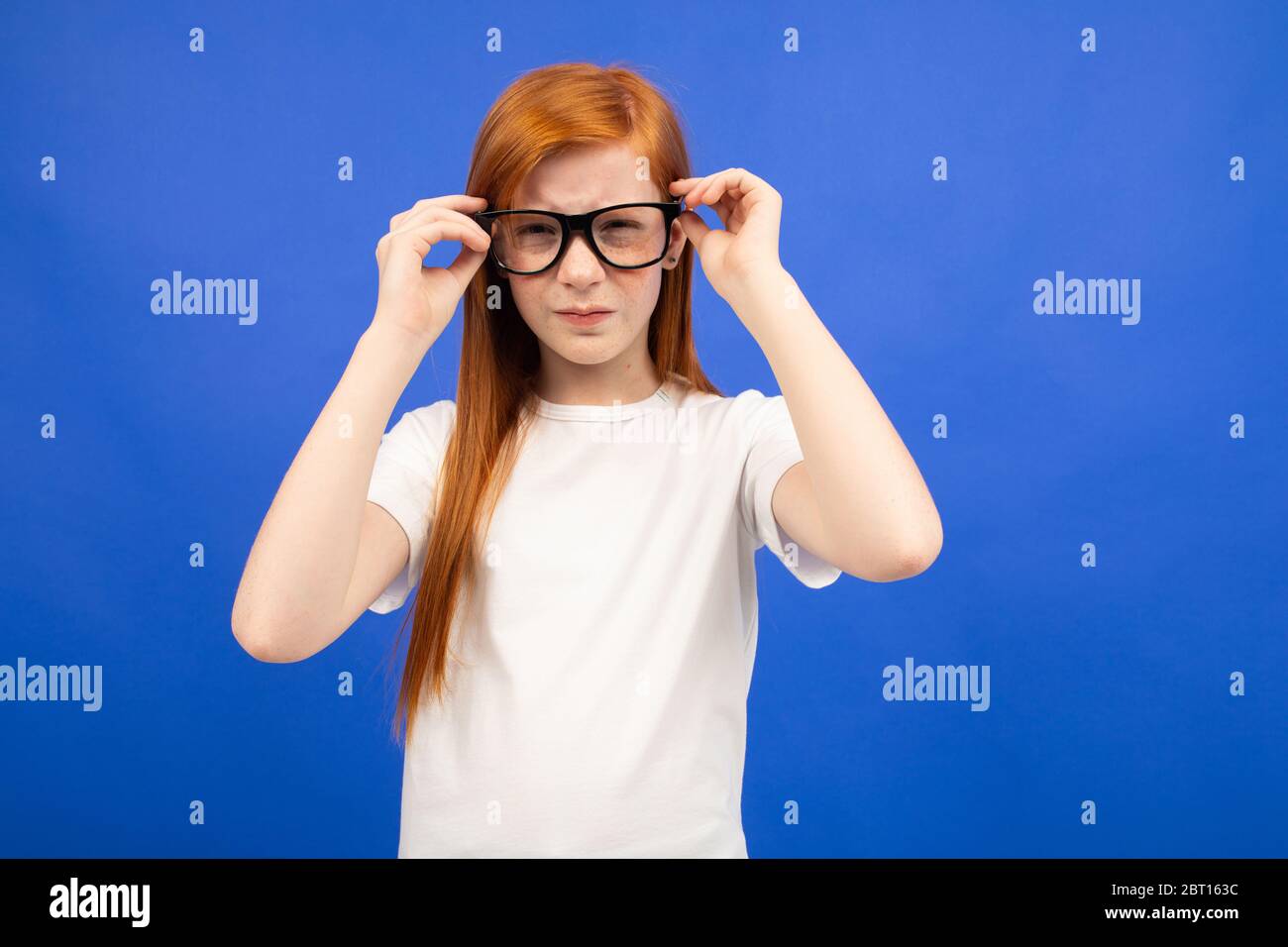 red-haired teenager girl squints while holding glasses in her hand on a blue studio background. Stock Photo