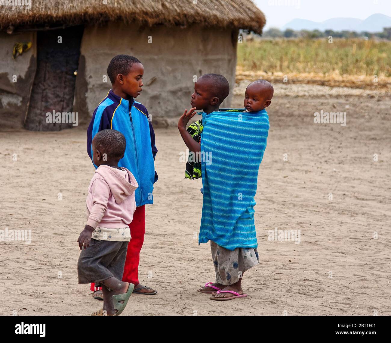 four Maasai children, village; indigenous people; small circular mud house, dirt, field, distant mountain, Tanzania; Africa Stock Photo