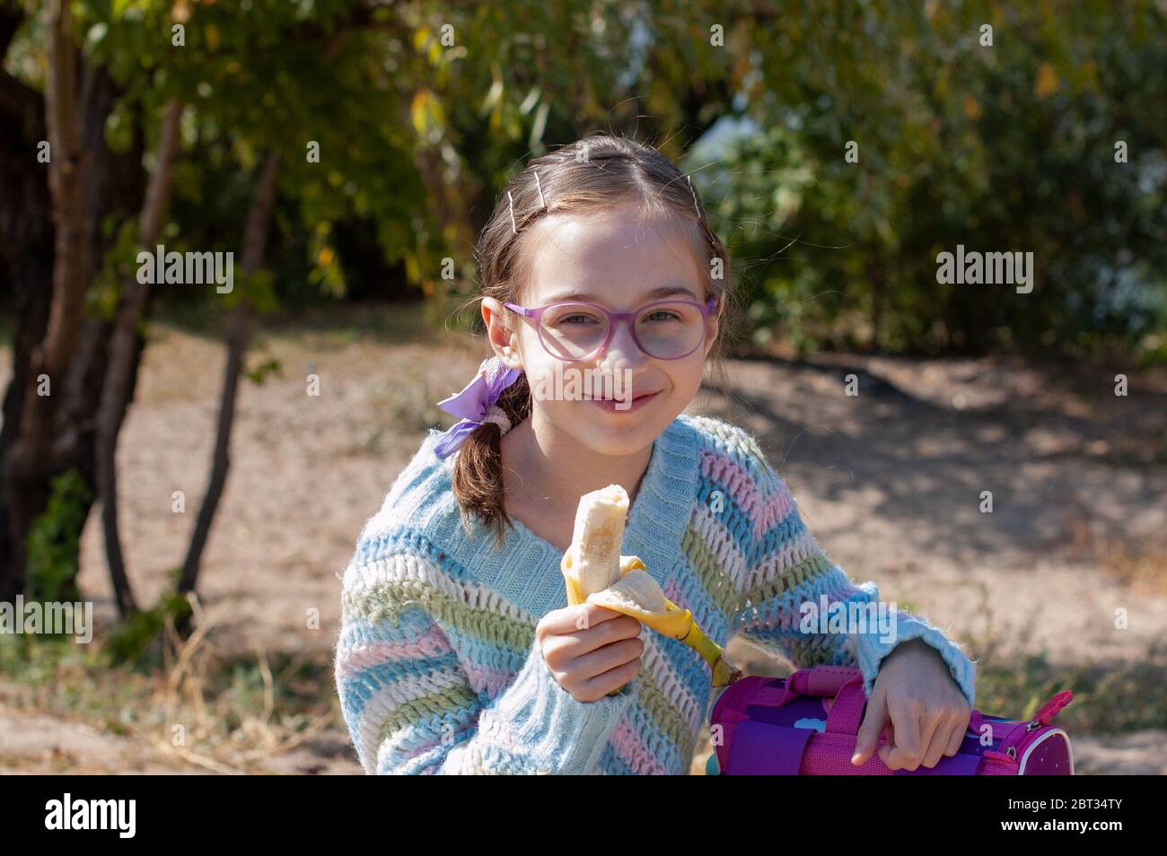 Schoolgirl eating a banana in the street. Alpha generation children will be the driving force behind progress in our century. They are more balanced, Stock Photo