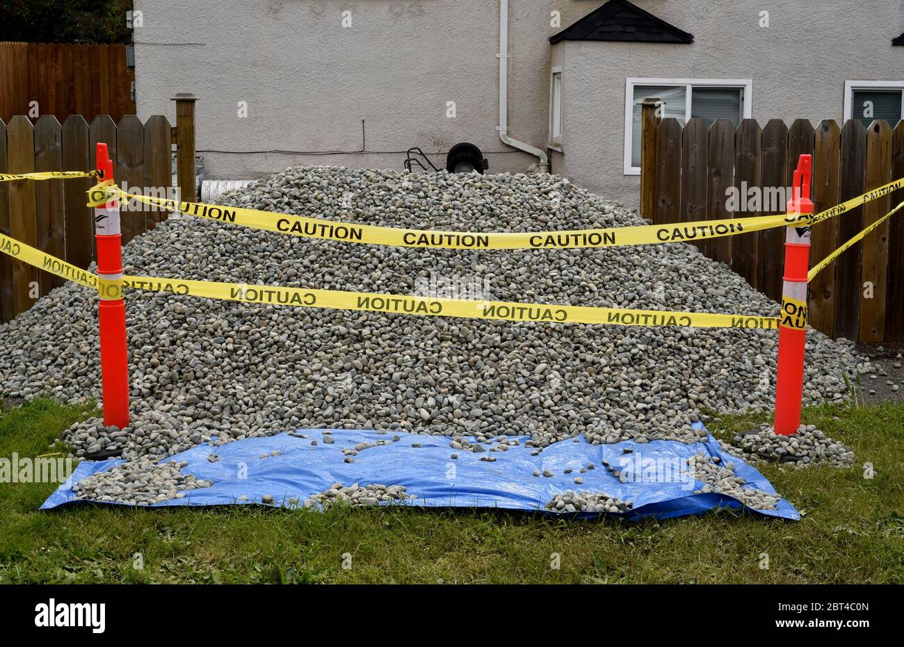 Caution tape surrounds a pile of gravel sittting on a blue tarp in front of a suburban house and yard. The small stones can be used for fill, drainage Stock Photo