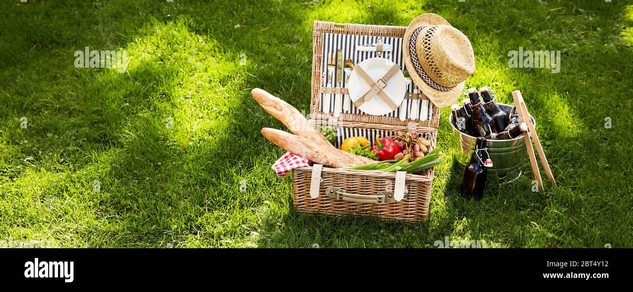 Vintage picnic hamper with vegetarian food and French baguettes adorned with an old straw hat alongside a silver cooler with bottle of beer on green g Stock Photo