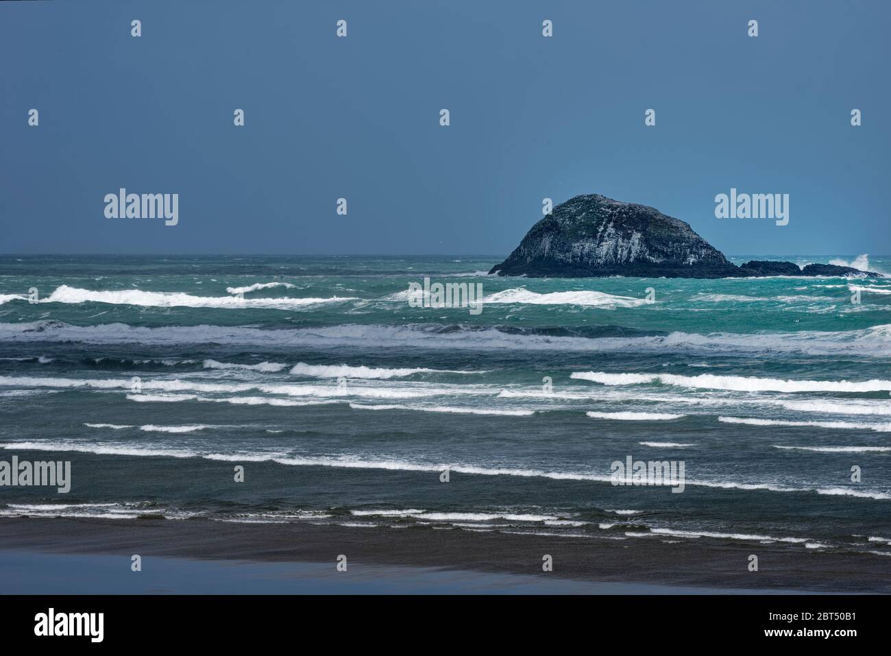 Surf on an empty beach on a windy day under clear skies and a rocky island in the background Stock Photo