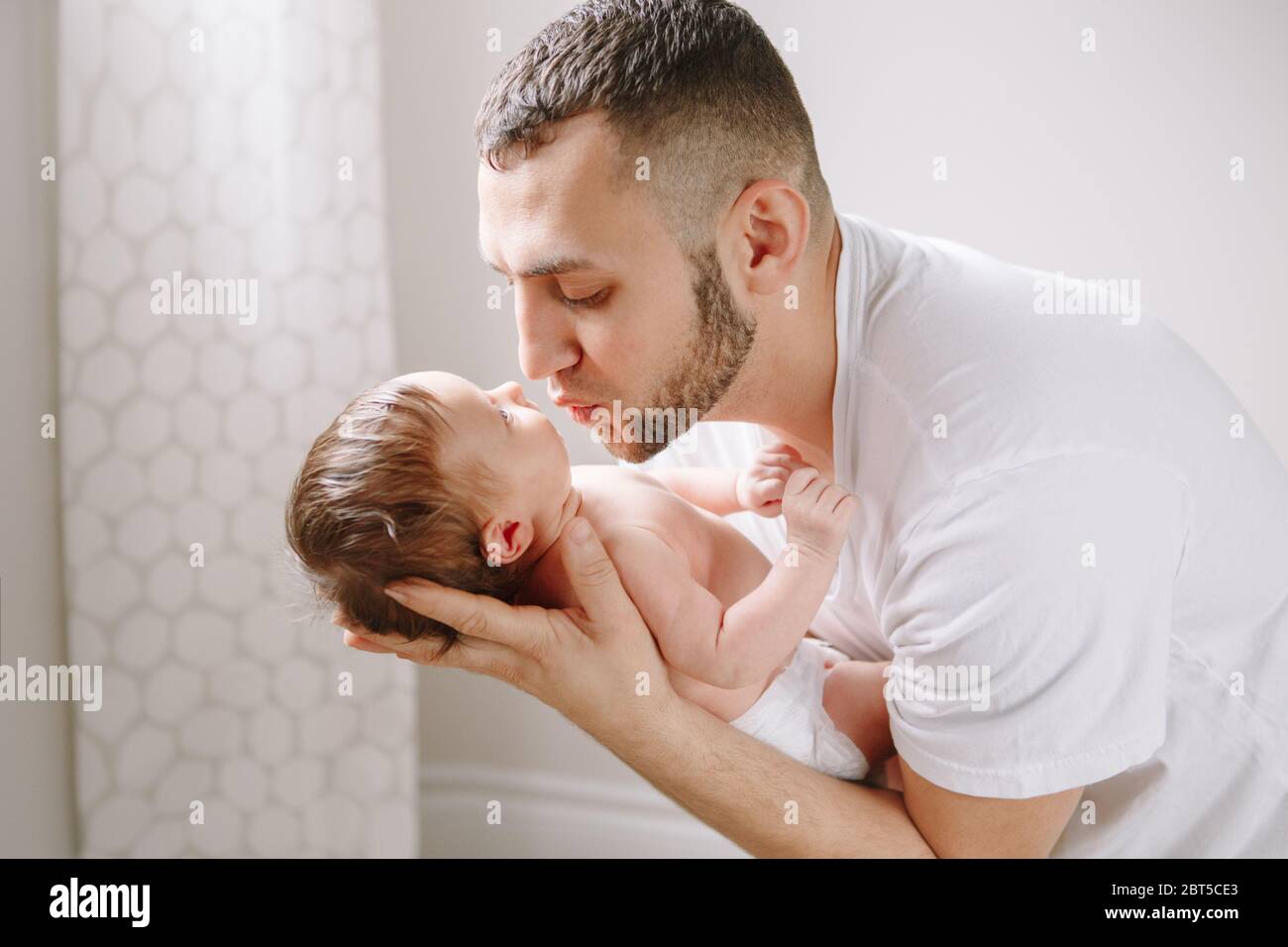 Happy young Caucasian father kissing newborn baby. Dad man parent holding rocking child daughter son on his hands. Authentic lifestyle candid family m Stock Photo