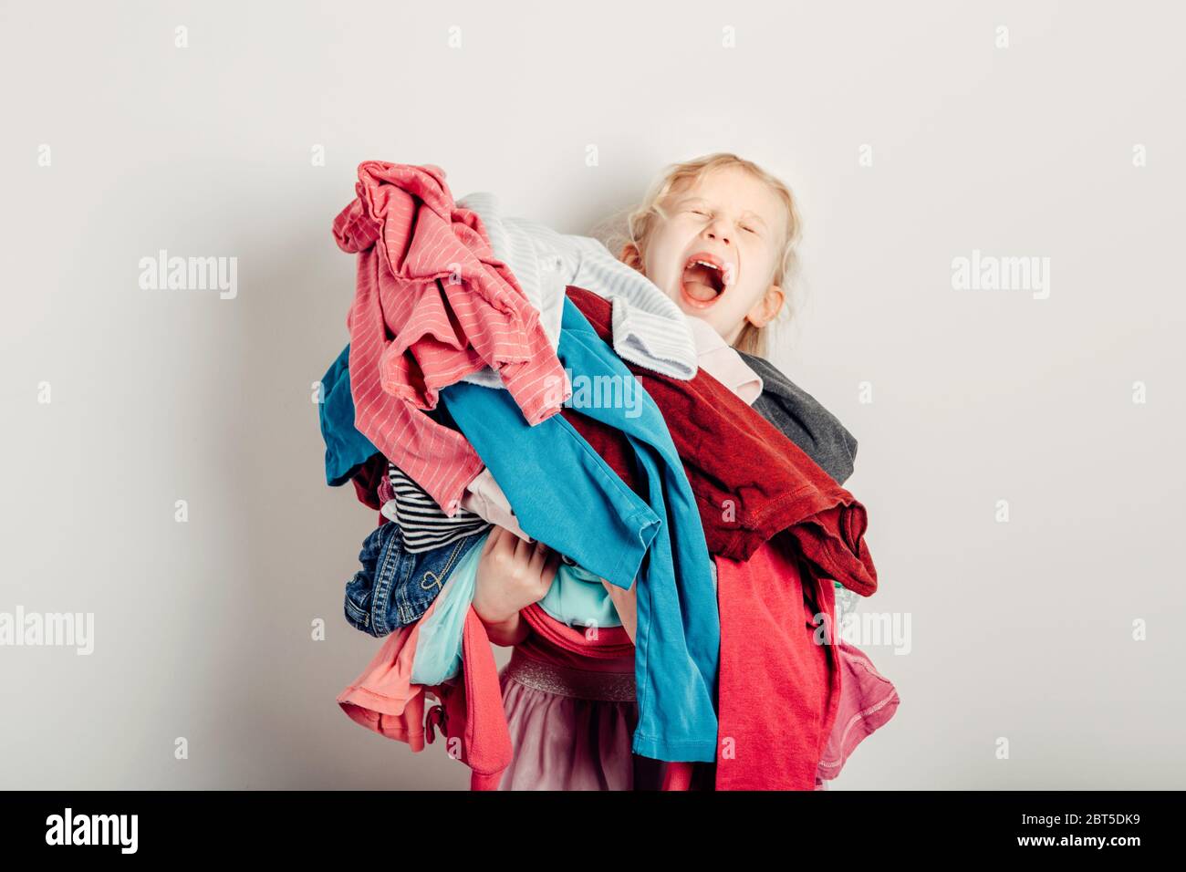 Mommy little helper. Cute Caucasian girl sorting clothes. Adorable funny child arranging organazing clothing. Kid holding messy stack pile of clothes Stock Photo