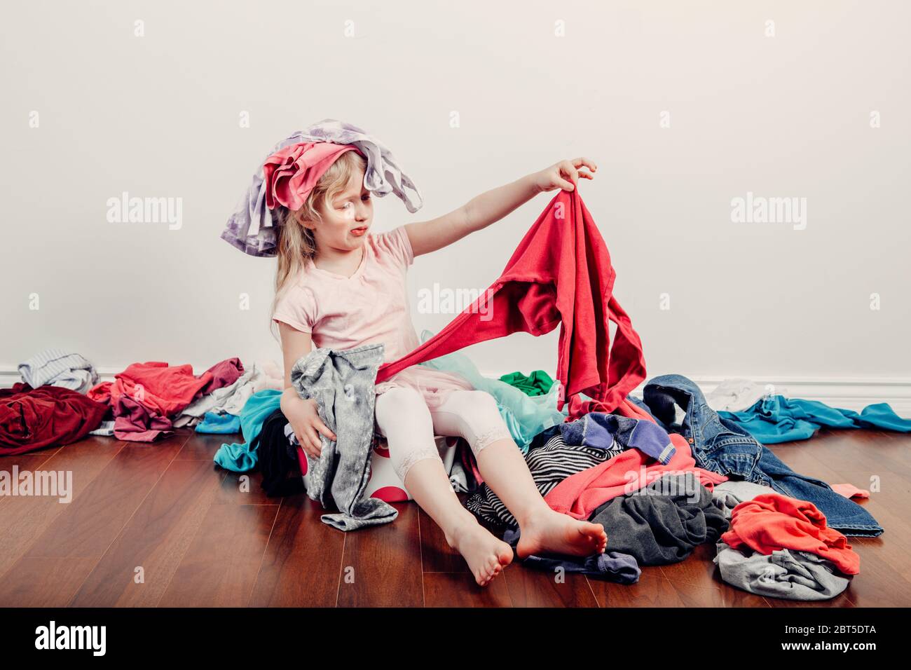 Kid playing with clothes on head. Cute Caucasian girl sorting clothes.  Adorable funny child arranging organazing clothing. Messy stack of clothes  thin Stock Photo - Alamy
